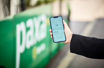 En hand håller fram en mobil som visar bokningssida i Paxa-appen. Paxa-buss i bakgrunden.